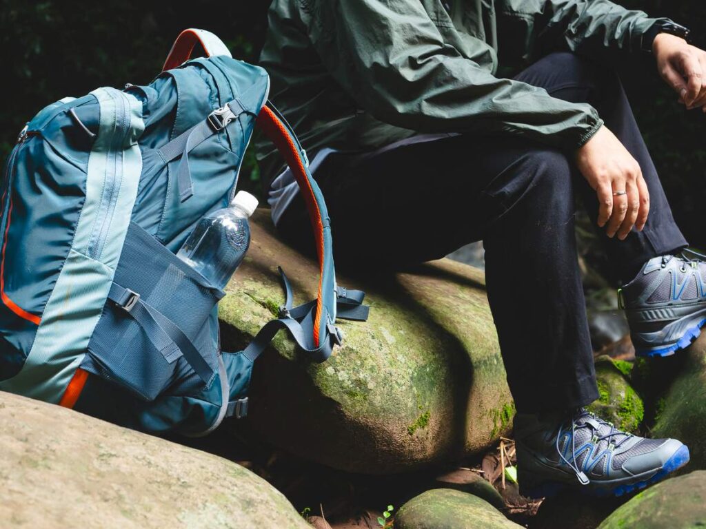 A hiker sitting on rocks in the forest wearing outdoor gear with a backpack and water bottle resting nearby.