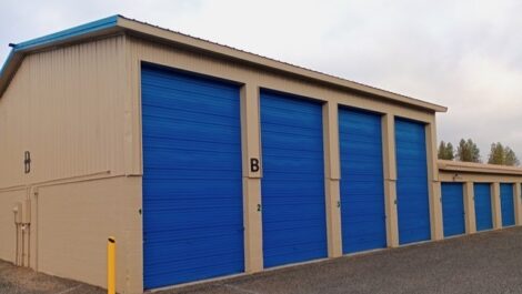 Beige metal storage building at Discount Mini Storage with bright blue roll-up doors along the side, including one tall blue door labeled 'B' with a yellow bollard in front, situated on a gravel or asphalt lot under an overcast sky.