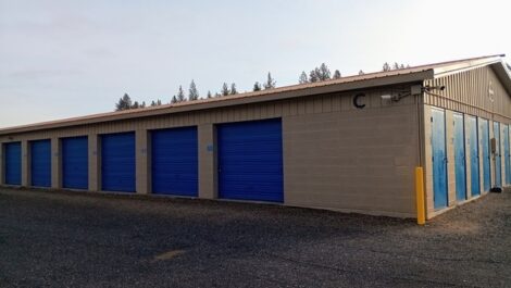 Beige metal storage building at Discount Mini Storage with bright blue roll-up doors along the side, including one tall blue door labeled 'B' with a yellow bollard in front, situated on a gravel or asphalt lot under an overcast sky.