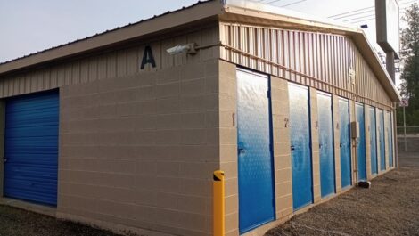Beige metal storage building at Discount Mini Storage with bright blue roll-up doors along the side, including one tall blue door labeled 'B' with a yellow bollard in front, situated on a gravel or asphalt lot under an overcast sky.