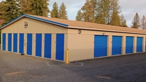 Beige metal storage building at Discount Mini Storage with bright blue roll-up doors along the side, including one tall blue door labeled 'B' with a yellow bollard in front, situated on a gravel or asphalt lot under an overcast sky.