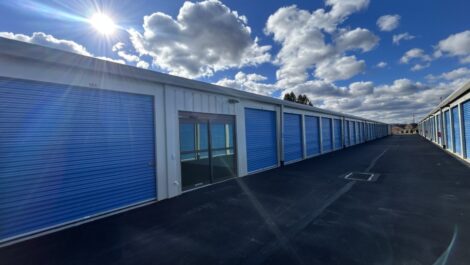 Beige metal storage building at State College with bright blue roll-up doors along the side, including one tall blue door labeled 'B' with a yellow bollard in front, situated on a gravel or asphalt lot under an overcast sky.