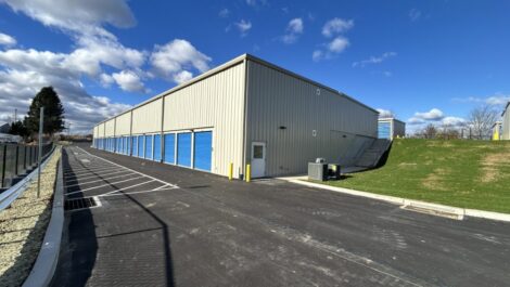 Exterior of State College Storage facility showing a fenced gravel lot with rows of tan metal storage buildings and roll-up doors, under a partly cloudy sky.