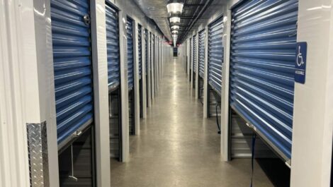 Row of green roll-up doors on tan metal storage units at State College Storage facility with gravel ground and trees behind.
