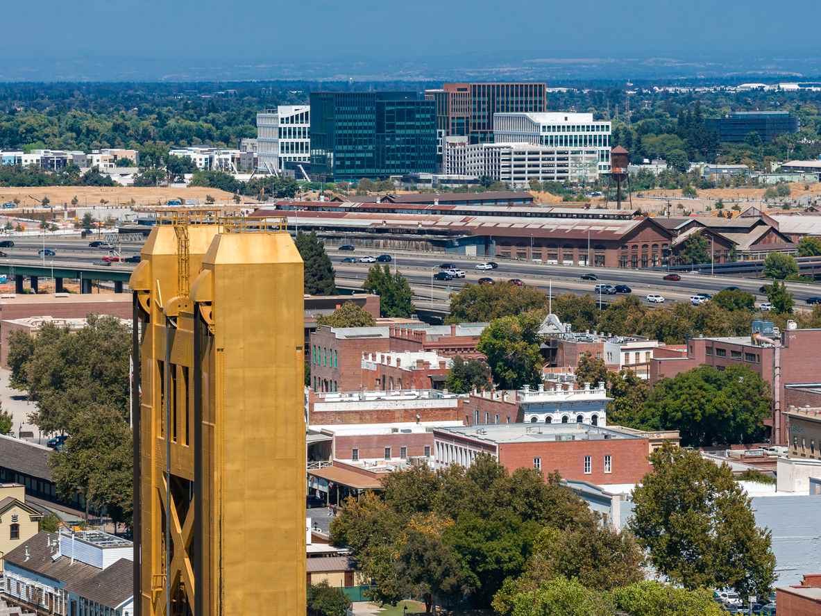 Sacramento, CA, featuring the gold Tower Bridge, downtown buildings, highways, and surrounding trees on a clear day.